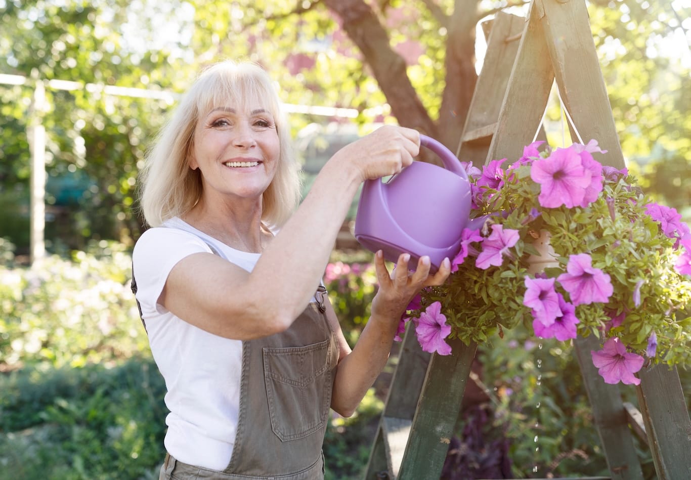 A woman in overalls waters a hanging basket of pink petunias with a purple watering can, enjoying the hobbies often pursued in dentistry for seniors and retirees.